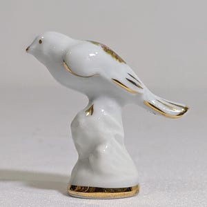 May include: A white porcelain bird figurine perched on a textured base. The bird features gold-painted trim along its wings, tail, and the base of the stand. A small, colourful floral decal is visible on the back of the bird, and the beak is gold.
