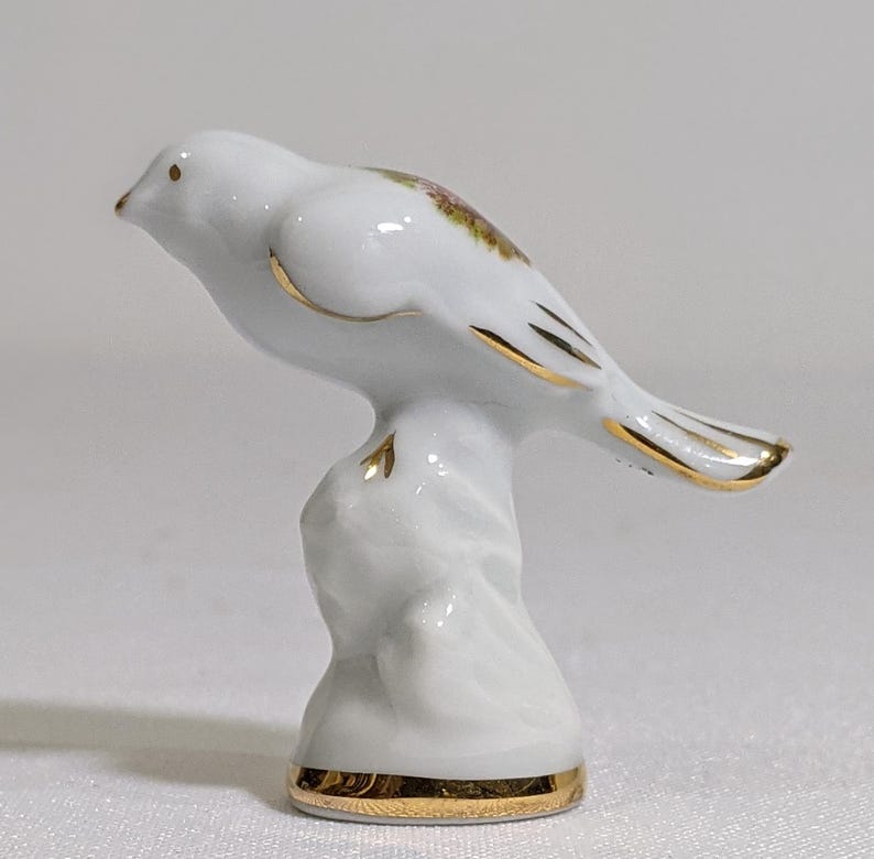 May include: A white porcelain bird figurine perched on a textured base. The bird features gold-painted trim along its wings, tail, and the base of the stand. A small, colourful floral decal is visible on the back of the bird, and the beak is gold.
