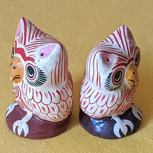 May include: Two small owl figurines featuring white bodies with intricate red line patterns, orange beaks, and black-outlined eyes. Each owl stands on a dark brown circular base with white claw details, showcasing a hand-painted aesthetic.