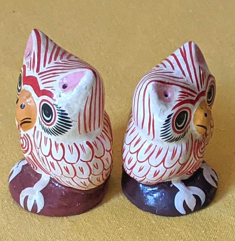 May include: Two small owl figurines featuring white bodies with intricate red line patterns, orange beaks, and black-outlined eyes. Each owl stands on a dark brown circular base with white claw details, showcasing a hand-painted aesthetic.