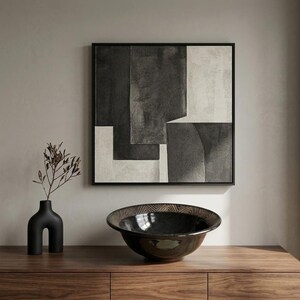 May include: A square framed wall art piece featuring an abstract composition of black, charcoal, and off-white geometric shapes. Beside it sits a matte black sculptural vase with a thin branch and a large, dark, wide-rimmed ceramic bowl.