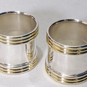 May include: Two silver napkin rings with gold-coloured bands. The rings have a cylindrical shape with a polished finish. The gold bands are evenly spaced around the circumference, adding elegance. Suitable for formal dining, these rings measure approximately 4 cm in diameter.