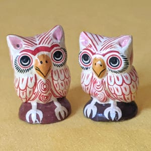 May include: Two small owl figurines featuring white bodies with intricate red line patterns, yellow beaks, and large black eyes. Each owl stands on a dark brown base with white claw details, showcasing hand-painted decorative elements.