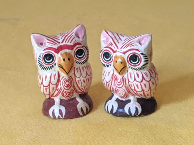 May include: Two small owl figurines featuring white bodies with intricate red line patterns, yellow beaks, and large black eyes. Each owl stands on a dark brown base with white claw details, showcasing hand-painted decorative elements.