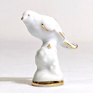 May include: A white porcelain bird figurine perched on a textured base. The bird features gold-painted accents on its beak, eye, wing edges, tail feathers, and feet. The base of the figurine is also trimmed with a band of metallic gold paint.