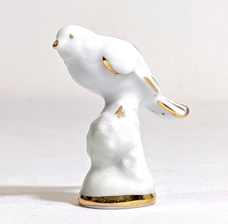 May include: A white porcelain bird figurine perched on a textured base. The bird features gold-painted accents on its beak, eye, wing edges, tail feathers, and feet. The base of the figurine is also trimmed with a band of metallic gold paint.
