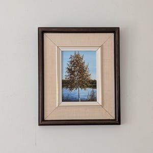 May include: A framed landscape painting depicting a tree with autumnal foliage, set against a blue sky and water. The artwork is enclosed in a dark brown wooden frame with a light beige mount, offering a classic aesthetic.