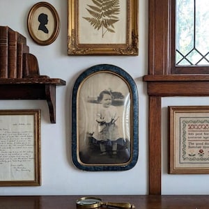 May include: A gallery wall arrangement featuring a central arched portrait of a child in a dark blue frame, surrounded by various framed prints, a handwritten letter, a cross-stitch sampler, and a brass magnifying glass resting on a surface.