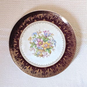 May include: A round decorative plate with a maroon border and gold filigree. The centre has a cream background with a floral arrangement in a gold vase. The plate has a gold rim and is suitable for display.