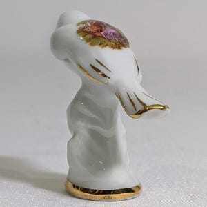 May include: A white porcelain thimble featuring a gold-trimmed base and gold accents on the top. The top surface displays a colourful, detailed transfer print of two figures in a garden setting. The thimble has a smooth, glossy finish throughout.
