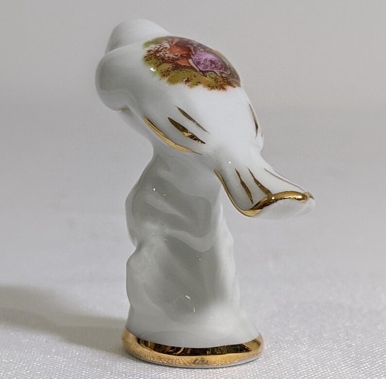 May include: A white porcelain thimble featuring a gold-trimmed base and gold accents on the top. The top surface displays a colourful, detailed transfer print of two figures in a garden setting. The thimble has a smooth, glossy finish throughout.