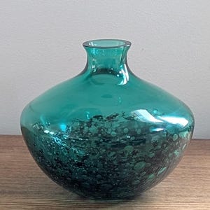 Puede incluir: Jarr&oacute;n de cristal color turquesa con cuerpo redondeado y cuello estrecho. La parte inferior del jarr&oacute;n presenta un dise&ntilde;o texturizado y moteado en tonos turquesa oscuro y negro. El jarr&oacute;n est&aacute; sobre una superficie de madera.