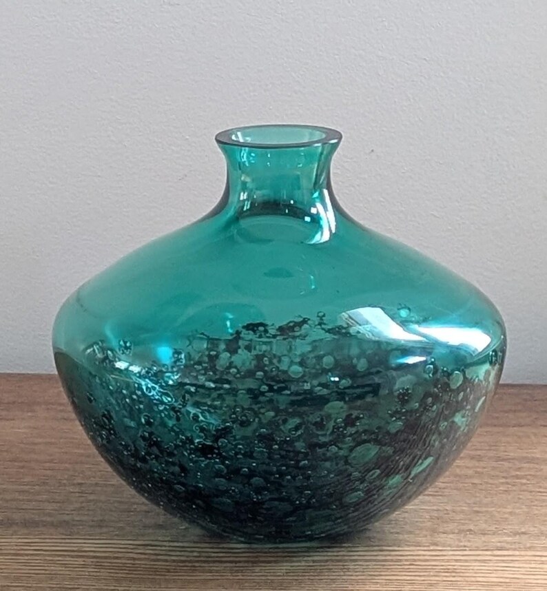 Puede incluir: Jarr&oacute;n de cristal color turquesa con cuerpo redondeado y cuello estrecho. La parte inferior del jarr&oacute;n presenta un dise&ntilde;o texturizado y moteado en tonos turquesa oscuro y negro. El jarr&oacute;n est&aacute; sobre una superficie de madera.