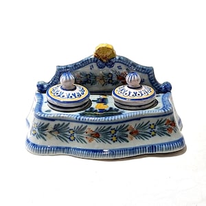 May include: A hand-painted ceramic inkwell with two ink pots and lids. The inkwell is decorated with blue, yellow, and white floral patterns. The ink pots have a rounded shape with a small knob on top. The inkwell is a decorative item.