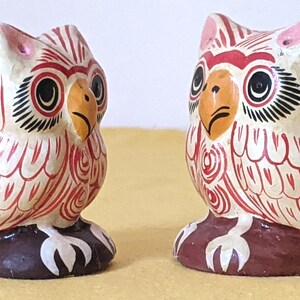 May include: Two small owl figurines crafted with a cream-colored base and detailed with intricate red line patterns. Each owl features a yellow beak, large black eyes with white accents, and pink ear tufts, all standing on a dark brown base.