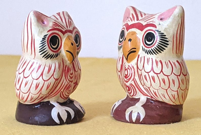 May include: Two small owl figurines crafted with a cream-colored base and detailed with intricate red line patterns. Each owl features a yellow beak, large black eyes with white accents, and pink ear tufts, all standing on a dark brown base.