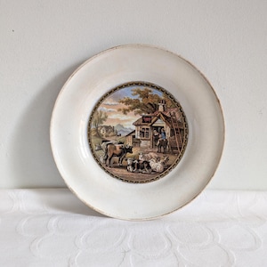 May include: A white ceramic plate featuring a detailed rural scene. The central image shows a cottage, cows, sheep, and a person on horseback. The plate has a slightly distressed, aged appearance, with a decorative border.
