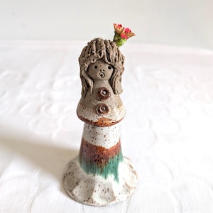 May include: A handcrafted ceramic vase, shaped like a girl, with a speckled finish. The vase has a brown head with curly hair, two brown buttons, and a flared base with brown and green stripes. A small pink flower sits on top of the head.