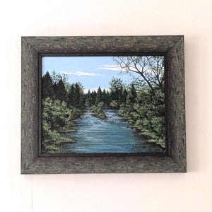 May include: Framed landscape painting showing a river winding through a forest. The artwork features a blue sky with clouds, green trees, and a dark grey textured frame. The painting captures a tranquil natural scene.