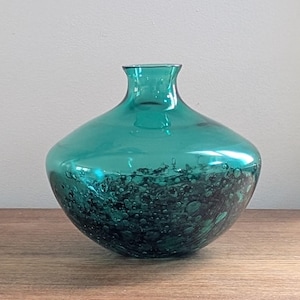 Puede incluir: Jarr&oacute;n de vidrio de color verde azulado con cuerpo redondeado y cuello estrecho. La parte inferior del jarr&oacute;n presenta un patr&oacute;n oscuro y texturizado, creando contraste con la parte superior lisa y transparente. El jarr&oacute;n est&aacute; sobre una superficie de madera.