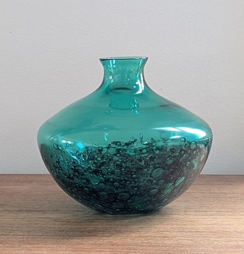 Puede incluir: Jarr&oacute;n de vidrio de color verde azulado con cuerpo redondeado y cuello estrecho. La parte inferior del jarr&oacute;n presenta un patr&oacute;n oscuro y texturizado, creando contraste con la parte superior lisa y transparente. El jarr&oacute;n est&aacute; sobre una superficie de madera.