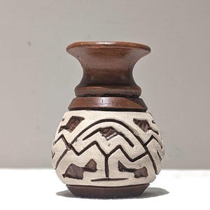 May include: A small, handcrafted vase with a dark brown top and base, and a cream-colored body. The body features a carved geometric design. The vase has a rounded shape with a narrow neck and a flared rim. The vase is likely made of ceramic or clay.