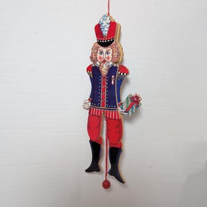 May include: A wooden nutcracker ornament with a red and blue uniform, a white hat with a blue feather, and a red and white striped sash. The nutcracker is holding a small gift box with a red bow. The ornament is hanging from a red string with a red ball at the end.