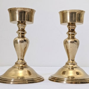 May include: A pair of gold metal candle holders with a classic design. Each holder has a round base, a tall, slender stem, and a wide, round cup at the top.