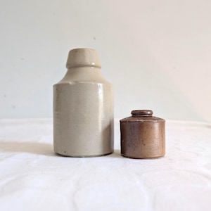 May include: Two antique stoneware bottles. The larger bottle is a light beige colour with a cylindrical body and a narrow neck. The smaller bottle is a dark brown colour with a rounded top. Both bottles have a rustic, aged appearance.
