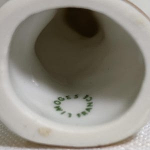 May include: A white porcelain bell with a hollow interior. The inner surface features a green circular stamp with the text LIMOGES FRANCE printed in a ring. The bell has a smooth, glossy finish and a wide, flared opening at the bottom edge.