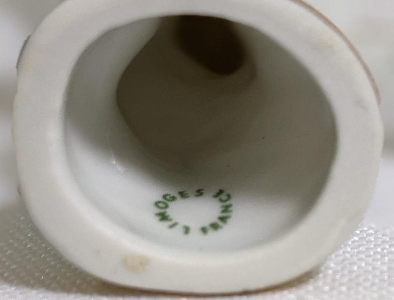 May include: A white porcelain bell with a hollow interior. The inner surface features a green circular stamp with the text LIMOGES FRANCE printed in a ring. The bell has a smooth, glossy finish and a wide, flared opening at the bottom edge.