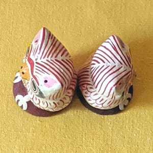 May include: Two small, hand-painted ceramic figurines shaped like birds, featuring white bodies with intricate red striped patterns, yellow beaks, and dark brown circular bases. The figurines are viewed from above, showing detailed painted feathers.