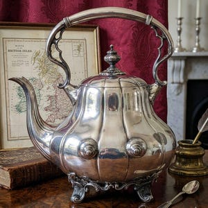 Puede incluir: Una tetera de plata pulida con cuerpo abombado y acanalado, pico curvo y un asa arqueada ornamentada. La tetera cuenta con una tapa extraíble con un pequeño remate y se apoya sobre tres patas decorativas con forma de voluta.