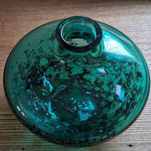 Puede incluir: Un jarr&oacute;n redondo de vidrio color turquesa, con un cuerpo ancho y un cuello estrecho. El vidrio tiene una apariencia burbujeante y texturizada, con patrones org&aacute;nicos oscuros. El jarr&oacute;n est&aacute; sobre una superficie de madera.