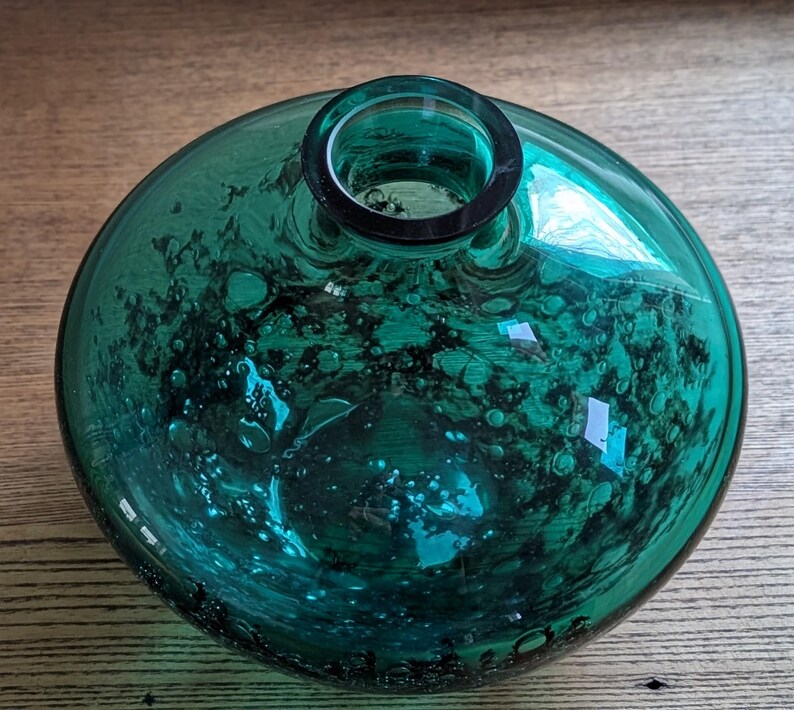Puede incluir: Un jarr&oacute;n redondo de vidrio color turquesa, con un cuerpo ancho y un cuello estrecho. El vidrio tiene una apariencia burbujeante y texturizada, con patrones org&aacute;nicos oscuros. El jarr&oacute;n est&aacute; sobre una superficie de madera.