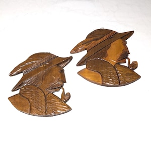 May include: Two wooden wall hangings depicting a man with a hat and pipe. The carvings are in shades of brown, with detailed textures and a rustic aesthetic. The man is seen in profile, with a hat and pipe.
