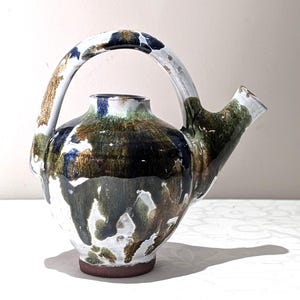 Peut inclure: Une théière en céramique faite à la main avec une anse et un bec courbés. La théière présente une base blanche avec des coulures de glaçure marron, verte et bleue. La anse est blanche avec des accents marron et bleu.