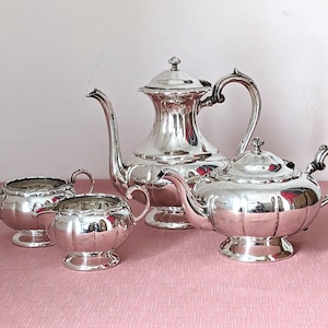 May include: A silver tea set comprising a teapot, coffee pot, sugar bowl, and creamer. The set is highly polished, reflecting the light. The teapot and coffee pot have curved spouts and handles. The set is displayed on a pink surface.
