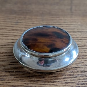 May include: A small, round, antique silver-toned container with a tortoiseshell top. The lid is a dark brown with lighter brown and black marbling. The container has a shiny, reflective surface and sits on a wooden surface.