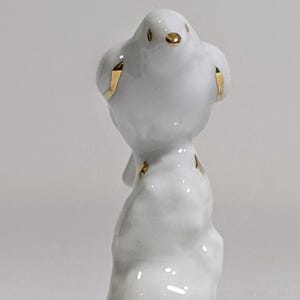 May include: A small white porcelain figurine of a bird perched on a textured base. The bird features delicate gold painted accents on its beak, eyes, wings, and tail feathers. A thin gold band encircles the bottom edge of the base.