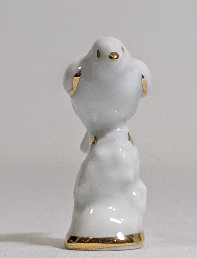 May include: A small white porcelain figurine of a bird perched on a textured base. The bird features delicate gold painted accents on its beak, eyes, wings, and tail feathers. A thin gold band encircles the bottom edge of the base.
