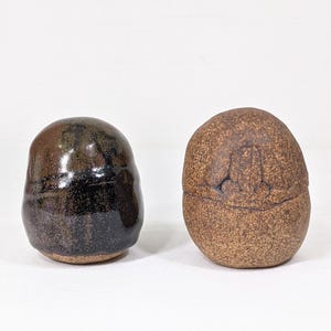 May include: Two ceramic, hand-thrown, round, lidded jars. One jar is brown with a rough texture. The other jar is black with a glossy finish.