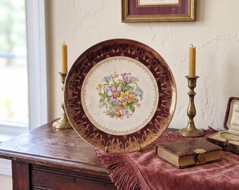 May include: A round ceramic plate featuring a central floral bouquet in shades of pink, yellow, and blue, surrounded by a wide, deep burgundy border with intricate gold filigree patterns and a scalloped edge, displayed on a small metal stand.