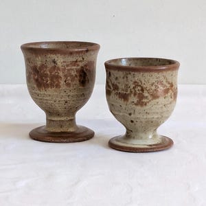 May include: Two handmade ceramic goblets. The goblets have a speckled beige and brown glaze with a flared rim and a pedestal base. The interior of the goblets is a darker brown. The goblets are of different sizes.