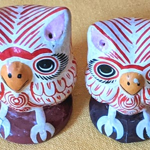 May include: Two hand-painted ceramic owl figurines featuring white bodies with intricate red line patterns, large black and white eyes, orange beaks, and white talons. Each owl is perched on a solid dark brown circular base, displaying a symmetrical design.
