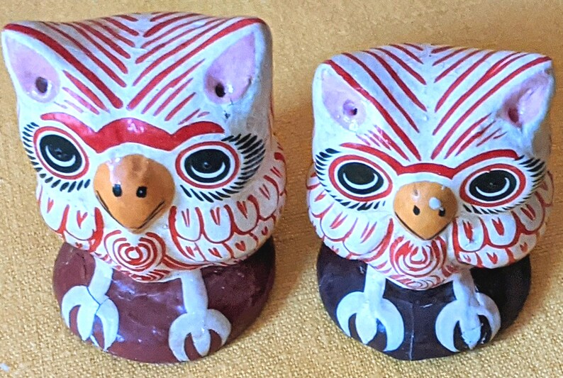 May include: Two hand-painted ceramic owl figurines featuring white bodies with intricate red line patterns, large black and white eyes, orange beaks, and white talons. Each owl is perched on a solid dark brown circular base, displaying a symmetrical design.