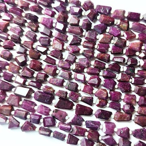 Può includere: Una collana di perle di rubino sfaccettate, ogni perla è di colore rosso intenso con una forma leggermente irregolare. Le perle sono infilate su un filo bianco.