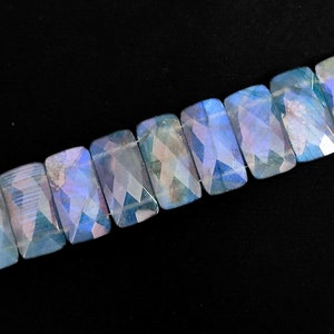 May include: A strand of iridescent blue and green labradorite gemstone beads, each cut into a rectangular shape with facets. The beads are strung on a dark cord.