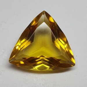 Pode incluir: Uma pedra preciosa triangular facetada com uma tonalidade amarela vibrante. A gema tem uma superfície polida e uma forma simétrica, refletindo a luz para criar uma exibição brilhante. A gema está sobre um fundo branco.