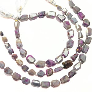 May include: Three strands of gemstone beads in shades of grey, purple, and brown. The beads are irregularly shaped and faceted. White tassels are attached to the ends of the strands, creating a layered effect.